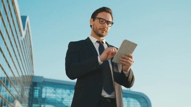 Attractive busineesman using digital tablet device while standing outside the office center building. Man reading, typing on tablet touchscreen and looking at the camera happy. Close up