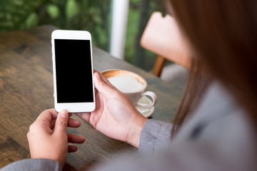 Mockup image of hands holding white mobile phone with blank white screen with hot coffee cup on wooden table in cafe