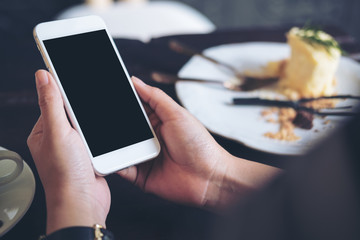 Mockup image of hands holding white mobile phone with blank black screen with a plate of cake on wooden table in restaurant