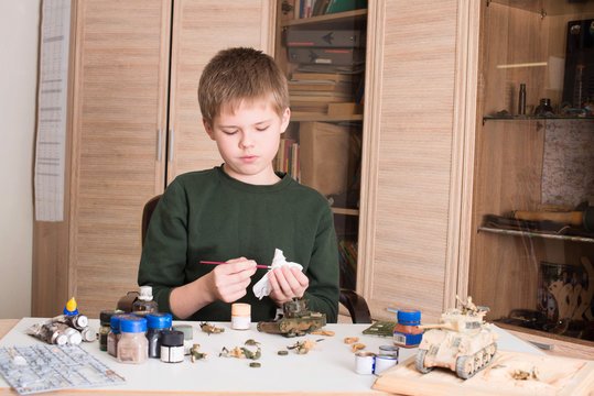 Hobby and leisure concept. Teen boy assembling and painting plastic model tank at workplace in his room.