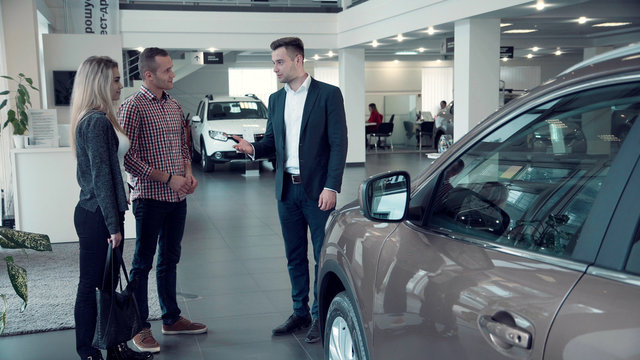 Young Salesman Delivering Sales Pitch To Couple Buying New Vehicle Inside Car Dealership While Standing In Front Of Shiny New Model Of Car Or Electrocar