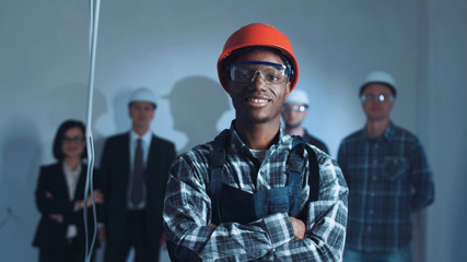 Handsome african young builder in protective form with hard hat standing and looking at camera on...