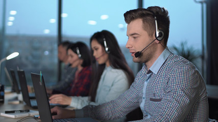 Young handsome men in checked shirt and headset working in call center in modern worldwide office...