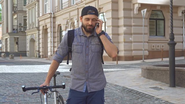 Handsome bearded cyclist using his cellphone on the street of the city. Attractive young man holding bicycle near him. Brunette caucasian guy dialing somebody on the phone