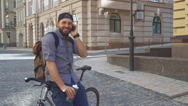Young brunette cyclist talks on the phone on the street of the city. Attractive bearded guy leaning on his bicycle frame. Handsome caucasian man with backpack holding his cellphone near his ear