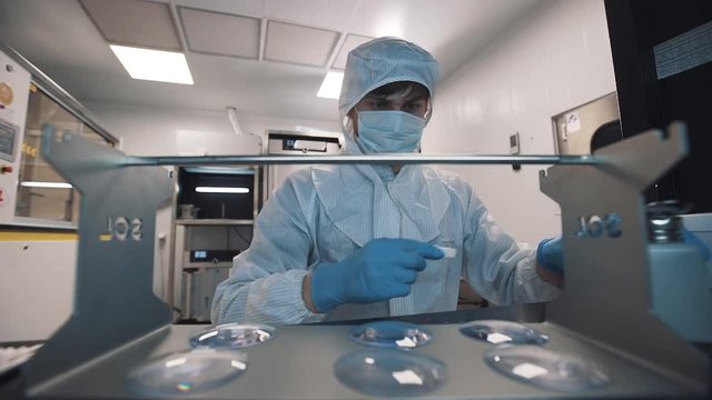Scientist caucasian man in sterile clothes, face mask and rubber gloves cleaning lenses using wet piece of cloth at laboratory