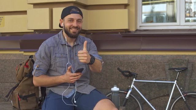 Attractive caucasian cyclist showing thumb up on the street. Handsome brunette guy approving biking in the city. Young bearded man listening to music on his smartphone