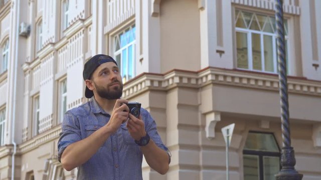 Handsome bearded cyclist taking a picture on the street of the city. Attractive brunette guy holding photographic camera. Young caucasian man in cap looking up against background of some public