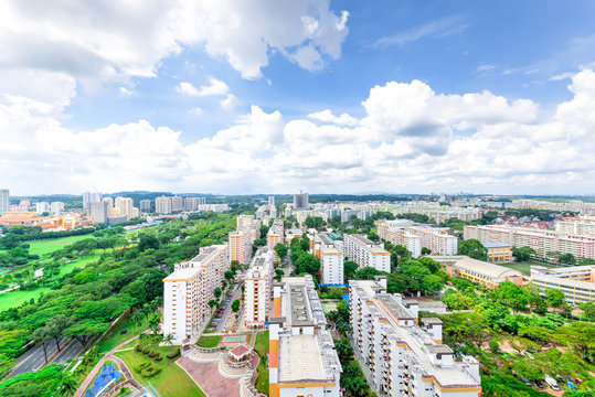Aerial view dense of HDB apartment buildings at midday in Ang Mo Kio neighborhood of Singapore. High rises flat complex with grass courtyard, green trees, tennis court, playground at blue cloud sky.