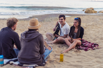 group of young and cheerful friends sitting on beach and lit bonfire. One man is playing guitar. Music on Wild beach