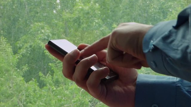 Closeup view of hands with black glossy smartphone. Caucasian man in blue shirt touch and scroll mobile phone screen. With blurred trees behind window at the back.