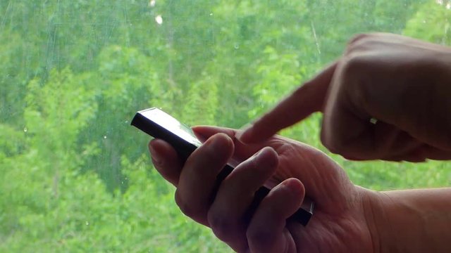 Closeup view of hands with black glossy smartphone. Caucasian man touch and scroll mobile phone screen. With blurred trees behind window at the back.