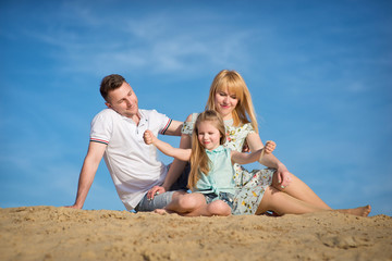 Happy beautiful laughing family sitting on the sand on the beach against the background blue sky in summer vacation.