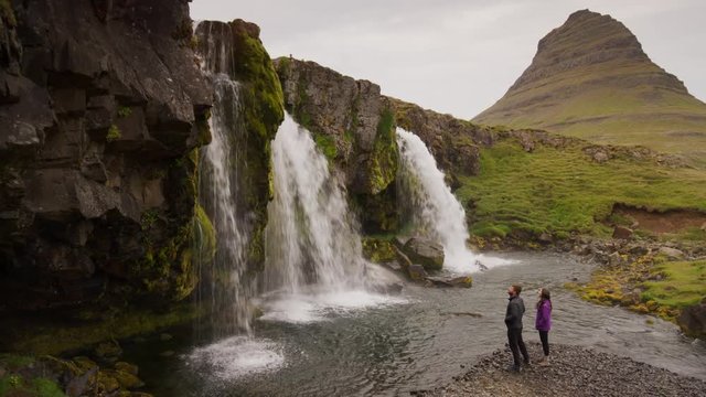 Wide panning shot of man photographing woman near waterfalls. Grundarfjorour, Snaefellsnes, Iceland