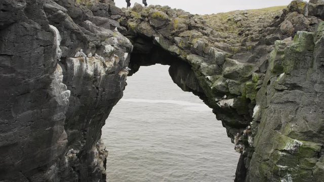 Wide crane shot of couple walking on arch rock formation. Arnarstapi, Snaefellsnes, Iceland