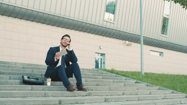 Young stylish businessman sitting on the stairs near office building, using digital tablet devide, talking on the cell phone and drinking coffee.