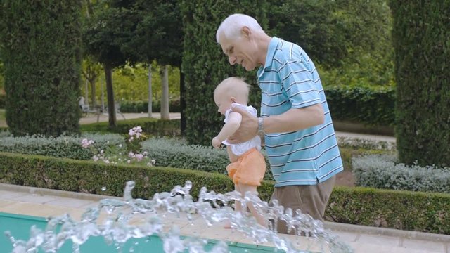 Baby learns to walk outdoors. Grandpa supports his granddaughter with both hands teaching to walk near fountain. Blonde baby girl takes first steps. Slow motion.