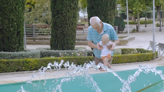 Baby learns to walk outdoors. Grandpa supports his granddaughter with both hands teaching to walk near fountain. Blonde baby girl takes first steps.