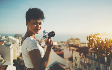Curly young Brazilian lady half turned towards camera and smiling while standing on high observation point in Lisbon city and holding vintage film camera with copy space for logo, message or advert