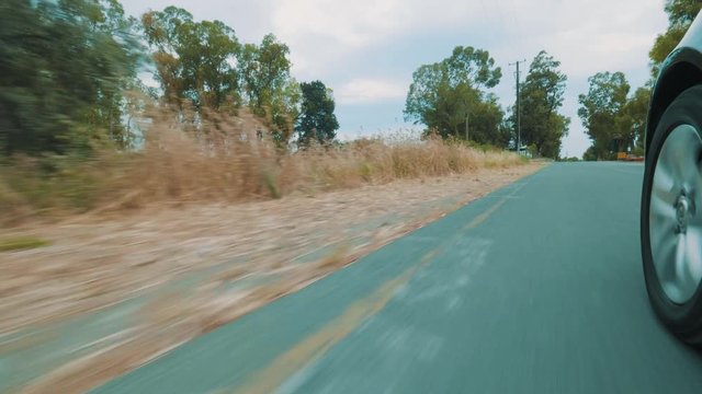 On the roadside are grass, trees and buildings. The car passes through haystacks, road signs. In front there is an even road and a green forest. View from on-board camera on weel and wing