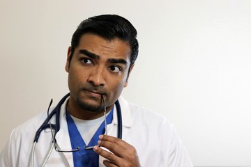 Young handsome doctor in blue scrubs thinking with glasses in mouth