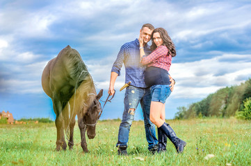 View through the grass: young couple - she is handsome brunette with long hair, pregnant; he is tall and brave, holding the reins of the black horse, a walk on the meadow. Wonderful sky behind them