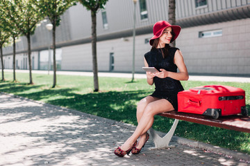 Woman in red hat checks her tablet sitting by a red suitcase