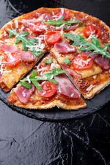 Homemade pizza with prosciutto, tomato, arugula on black slate board. Close up.