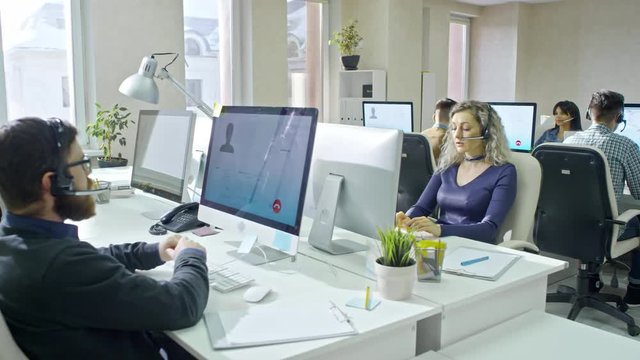 PAN of male and female call centre operators in headsets sitting before computers in airy office and talking to clients via online voice calls 
