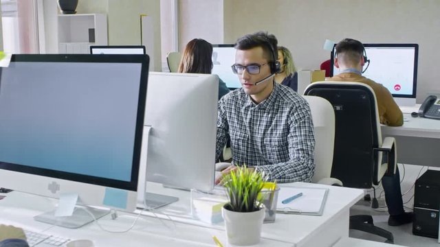 PAN of call centre operator in glasses and his male and female colleagues sitting in airy office and answering voice calls from customers on computers 