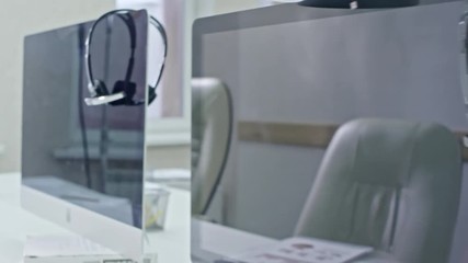 PAN with close up of computer monitors with headsets hanging on them standing on desks in empty office 