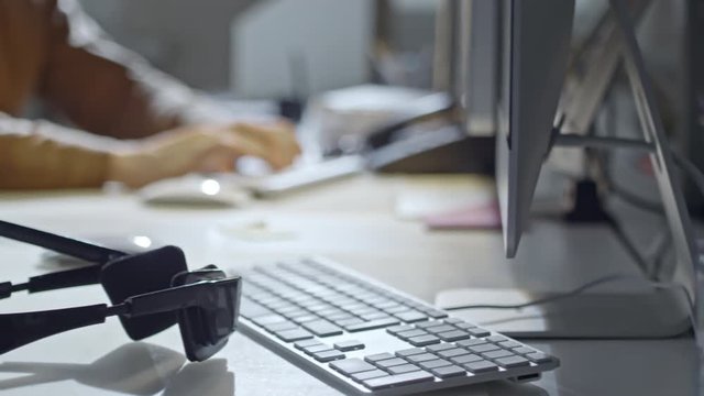 PAN of headphones with microphone lying before computer on office desk; blurred unrecognizable person working in background 