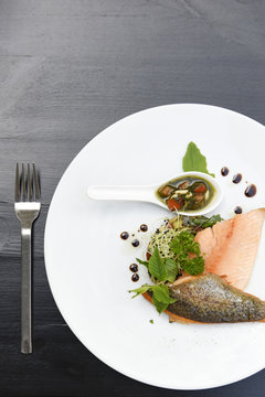 Plate of salmon fillets with fresh herbs, vegetables and balsamic vinegar on a black table
