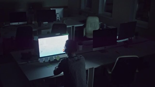 Tilt down crane shot of man wearing headset with microphone and sitting on exercise ball before computer in dark empty office while talking on voice call