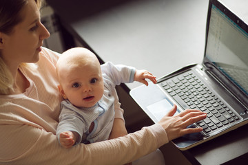 Young beautiful blonde woman, mother in home office with computer and her baby. Work and child care.
