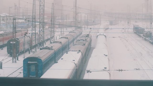 Passing by rail road depo full of trains and hight voltage lines covered in snow, winter cold blizzard day, slow motion