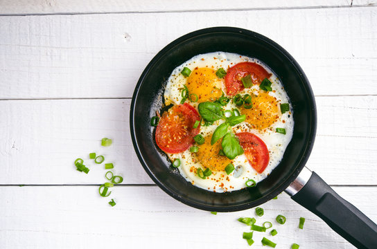 Pan of fried eggs with tomatoes, cheese, spring onion, herbs on a white table. White wooden table. Concept of food. Breakfast time. Copy space. 