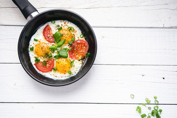 Pan of fried eggs with tomatoes, cheese, spring onion, herbs on a white table. White wooden table. Concept of food. Breakfast time. Copy space. 