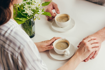 cropped shot of young couple drinking coffee and holding hands while sitting at table