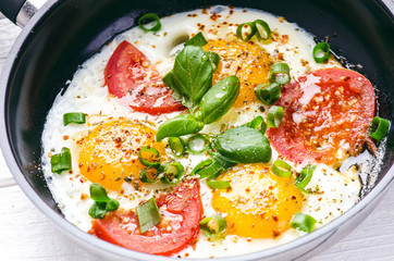 Pan of fried eggs with tomatoes, cheese, spring onion, herbs on a white table. White wooden table. Concept of food. Breakfast time. Copy space. 