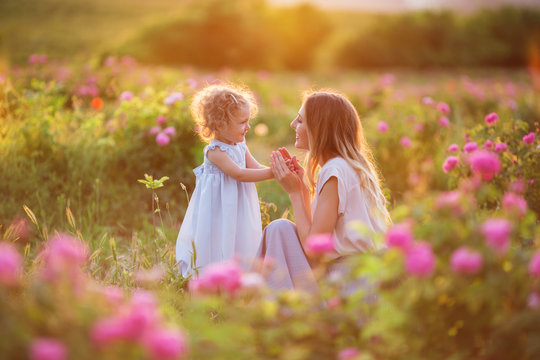 Beautiful child girl with young happy mother are wearing casual clothes walking in roses garden over sunset lights, summer time