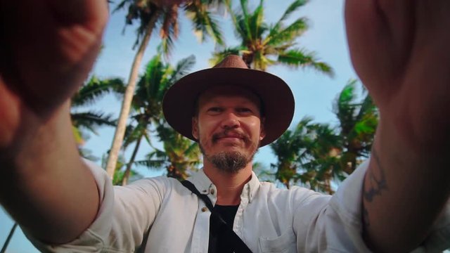 A young man in a hat with moustache among coconut palms keeps the camera or phone in your hands, shoots himself and sends greetings in the camera