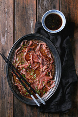 Asian style Beef carpaccio with soy sauce, chive onion, black sesame, served on vintage metal tray with textile, bowl of sauce and chopsticks over old wooden plank background. Top view with space