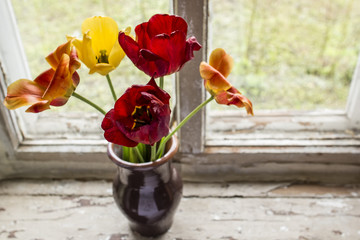 Beautiful blooming tulips in a clay vase standing at the window on an old windowsill with cracked paint.