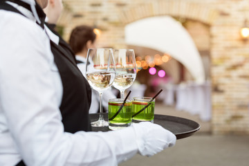 Midsection of professional waiters in uniform serving wine, cocktails and snacks during buffet catering party, festive event or wedding. Full glasses of wine on tray. Outdoor party catering service.