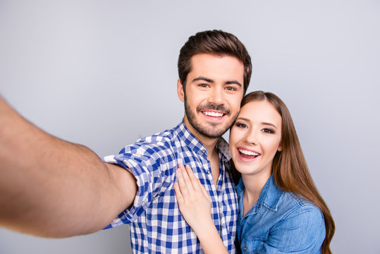 Close up of a cheerful young couple making selfie photo on guy`s camera. They are in casual outfits, posing and smiling on pure light background, embracing