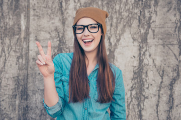 Close up portrait of cheerful playful girl, who is gesturing peace sign and smiles. She is wearing casual clothes, glasses and a brown hat, walking outdoors near concrete wall