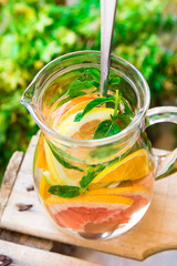 Glass pitcher with detox infused citrus water with grapefruits, oranges, limes, lemons, fresh mint, wooden garden fruit box, green plants, authentic atmosphere, cleansing