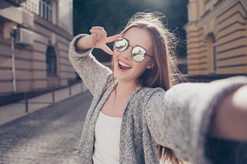 Carefree and happy, sunny spring mood. Cute young smiling girl is making selfie on a camera while walking outdoors. She is wearing casual outfit, mirror glasses