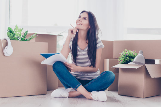 Cute teen in casual clothes is sitting with crossed legs on the floor. A lot of unpacked boxes near her. She is thinking and making notes in her diary